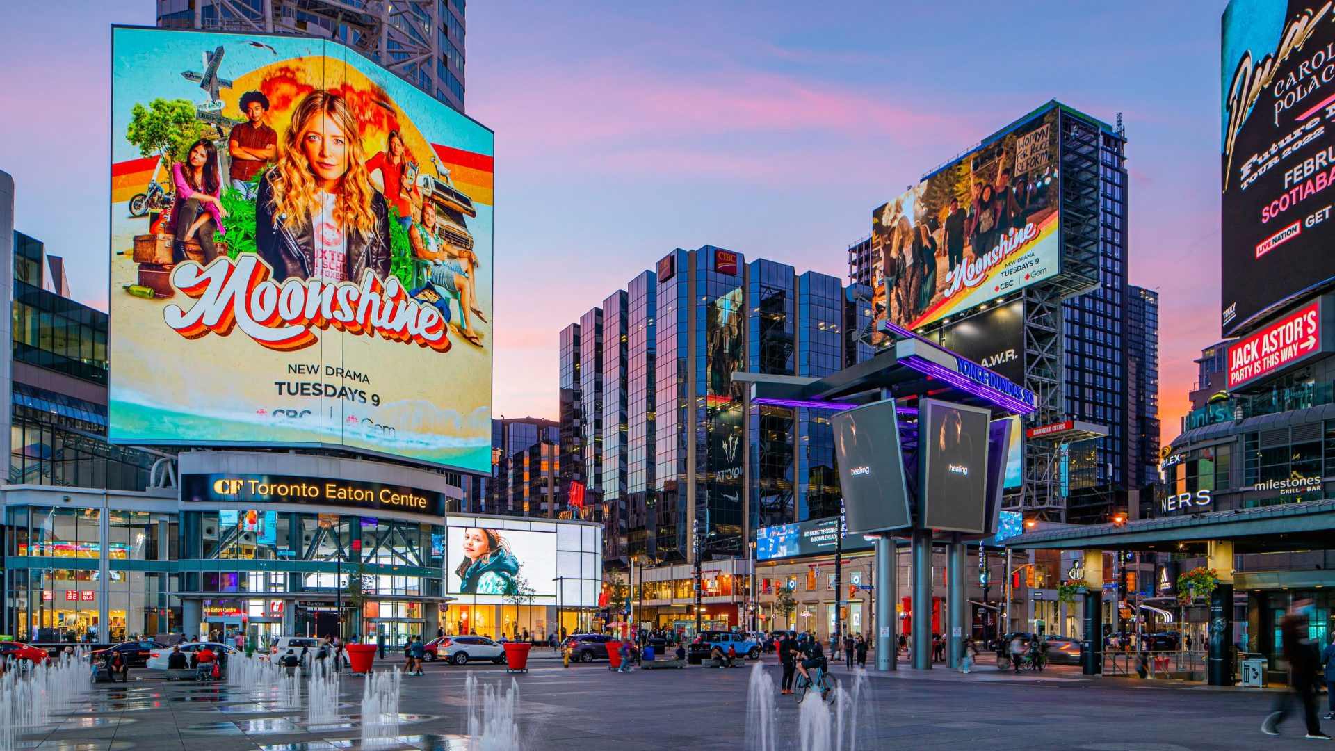 Yonge-Dundas Square in Toronto at sunset, featuring large digital billboards including a prominent ad for the TV drama ‘Moonshine,’ with people gathered near the fountains and modern buildings in the background.