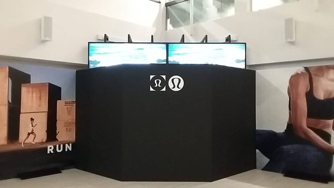 Lululemon trade show display with branded black kiosk, large wall graphics, and dual overhead digital screens.