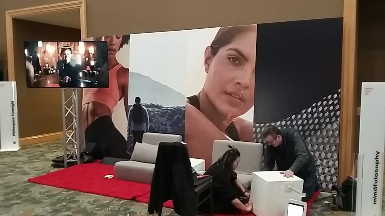 Trade show booth with large lifestyle wall graphics, lounge seating, and a mounted video display screen showcasing brand content.