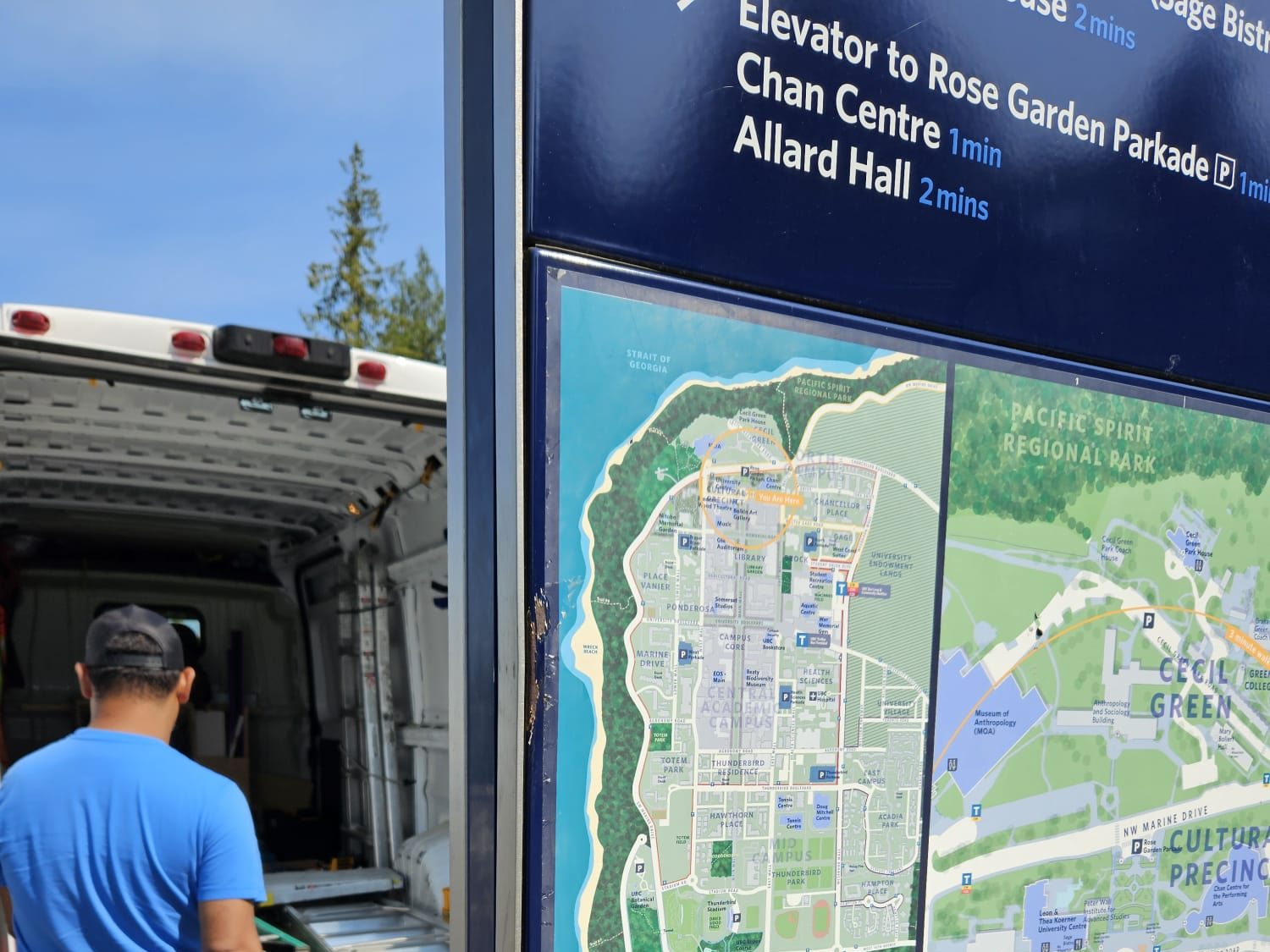 A close-up, outdoor shot of a blue wayfinding sign at the University of British Columbia (UBC). The top section features directional text with walking times: "Elevator to Rose Garden Parkade 1 min," "Chan Centre 1 min," and "Allard Hall 2 mins." Below, a detailed campus map highlights the "Campus Core," "Pacific Spirit Regional Park," and the "Cultural Precinct." A person in a blue shirt stands to the left near a white van, with trees and sky in the background.